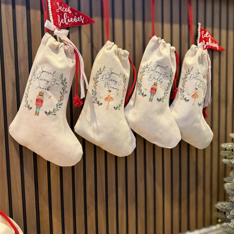 May include: Four white canvas stockings with red drawstrings hanging on a wood wall. Each stocking has a different design with a wreath and a character in the center. The stockings say "Special Delivery For" followed by a name. The names are Jack, Manny, Delia, and Ella.