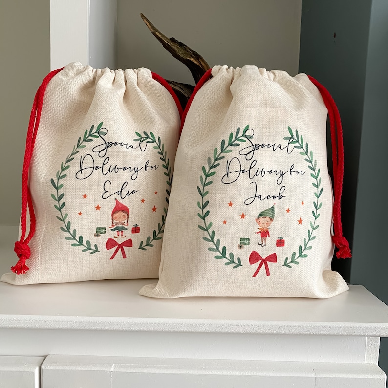 May include: Two white fabric gift bags with red drawstrings. Each bag has a green wreath with red berries and stars. The wreath surrounds the text "Special Delivery for" and a name. One bag has a girl elf illustration and the other has a boy elf illustration.