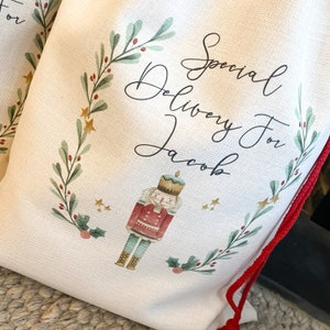 May include: A white canvas Christmas gift bag with a red drawstring. The bag features a green wreath with red berries and gold stars. The text "Special Delivery For Jacob" is written in black cursive script. A cartoon nutcracker is in the center of the bag.