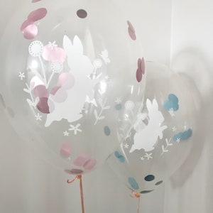 May include: Two clear balloons with white bunny silhouettes and confetti inside. The balloon on the left has pink and brown confetti, while the balloon on the right has blue and black confetti.