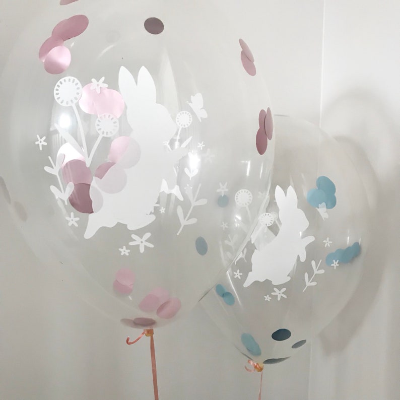 May include: Two clear balloons with white bunny silhouettes and confetti inside. The balloon on the left has pink and brown confetti, while the balloon on the right has blue and black confetti.