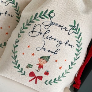 May include: A white canvas bag with a red drawstring. The bag has a green wreath design with the text "Special Delivery for Jacob" and an illustration of an elf carrying gifts.