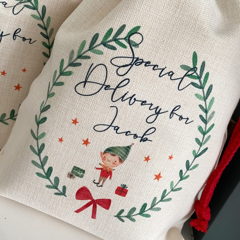 May include: A white canvas bag with a red drawstring. The bag has a green wreath design with the text "Special Delivery for Jacob" and an illustration of an elf carrying gifts.
