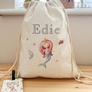May include: A personalised drawstring bag with a mermaid design and the name "Edie" embroidered on the front. The bag is made of natural canvas and has a matching wooden tag that says "Edie's PE Kit".