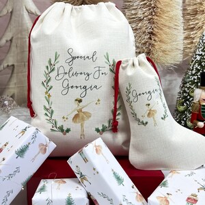 May include: Two white fabric gift bags with red drawstrings. The larger bag has the text "Special Delivery For Georgia" and a ballerina illustration. The smaller bag has the text "Georgia" and a ballerina illustration. Both bags are decorated with green leaves and gold stars. The bags are sitting on a red surface with several wrapped gifts.