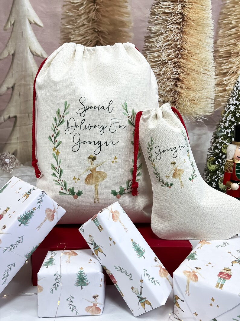 May include: Two white fabric gift bags with red drawstrings. The larger bag has the text "Special Delivery For Georgia" and a ballerina illustration. The smaller bag has the text "Georgia" and a ballerina illustration. Both bags are decorated with green leaves and gold stars. The bags are sitting on a red surface with several wrapped gifts.