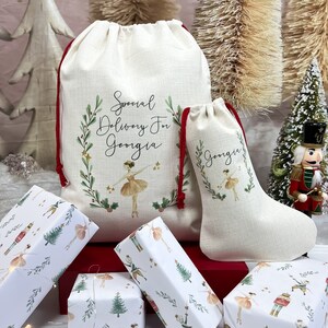 May include: Two white fabric gift bags with red drawstrings. The larger bag has the text "Special Delivery For Georgia" with a green wreath and a ballerina illustration. The smaller bag has the text "Georgia" with a green wreath and a ballerina illustration. Both bags are sitting on a red surface with wrapped gifts.