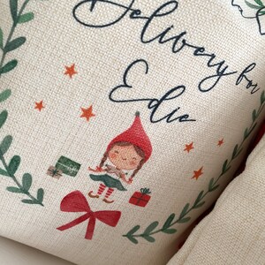 May include: A beige fabric pillow with a green wreath border, a red bow, and a cartoon elf holding a gift. The pillow has the text "Delivery for Edie" printed on it.