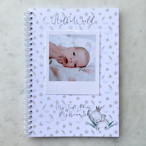 May include: A white baby book with a spiral binding. The cover features a gray and white star and crescent moon pattern. The text "Hello World" is written in cursive. A photo of a baby is in a white frame on the cover. The text "My First Photo My Name Is" is written in cursive. A cartoon elephant with a crown is on the cover.