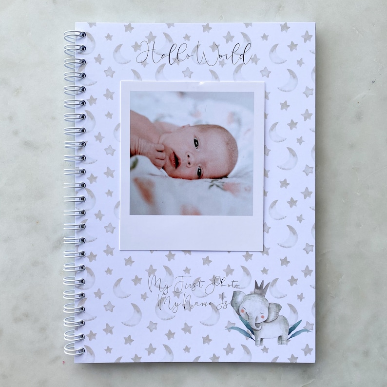 May include: A white baby book with a spiral binding. The cover features a gray and white star and crescent moon pattern. The text "Hello World" is written in cursive. A photo of a baby is in a white frame on the cover. The text "My First Photo My Name Is" is written in cursive. A cartoon elephant with a crown is on the cover.