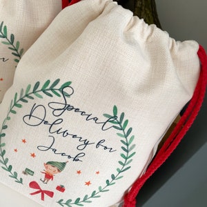 May include: A white canvas gift bag with a red drawstring. The bag has a green wreath with the text "Special Delivery for Jacob" and a cartoon elf holding a gift.