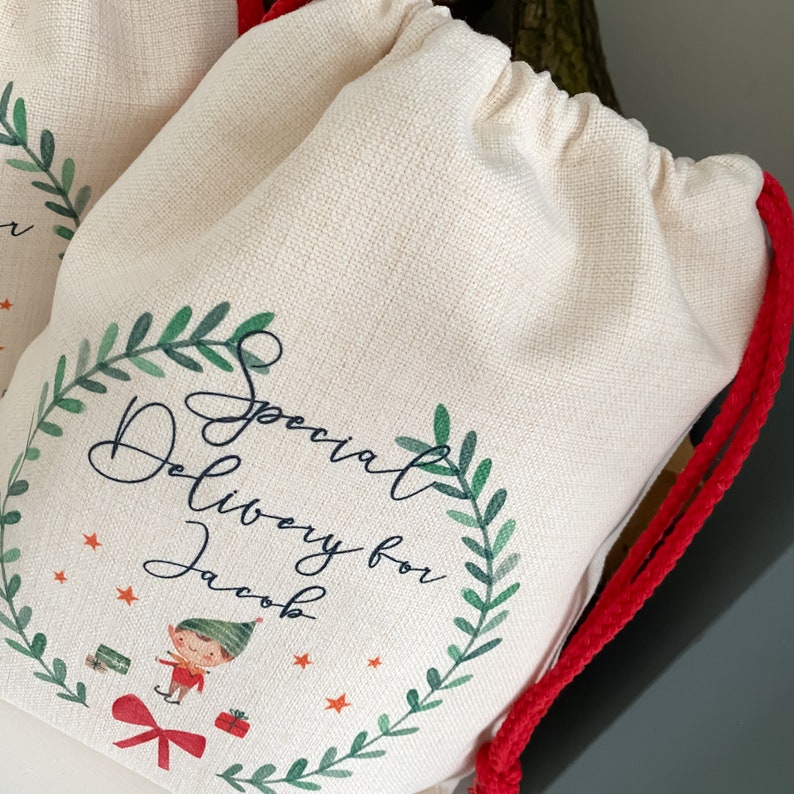 May include: A white canvas gift bag with a red drawstring. The bag has a green wreath with the text "Special Delivery for Jacob" and a cartoon elf holding a gift.