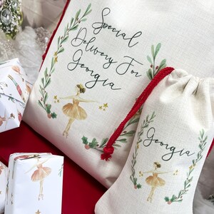May include: Two white canvas gift bags with red drawstrings. The larger bag has a ballerina illustration and the text "Special Delivery For Georgia". The smaller bag has a ballerina illustration and the text "Georgia". Both bags have a green leafy border.