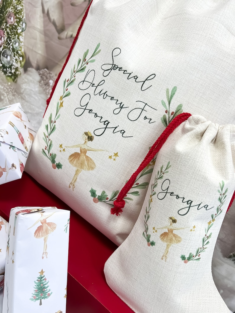 May include: Two white canvas gift bags with red drawstrings. The larger bag has a ballerina illustration and the text "Special Delivery For Georgia". The smaller bag has a ballerina illustration and the text "Georgia". Both bags have a green leafy border.