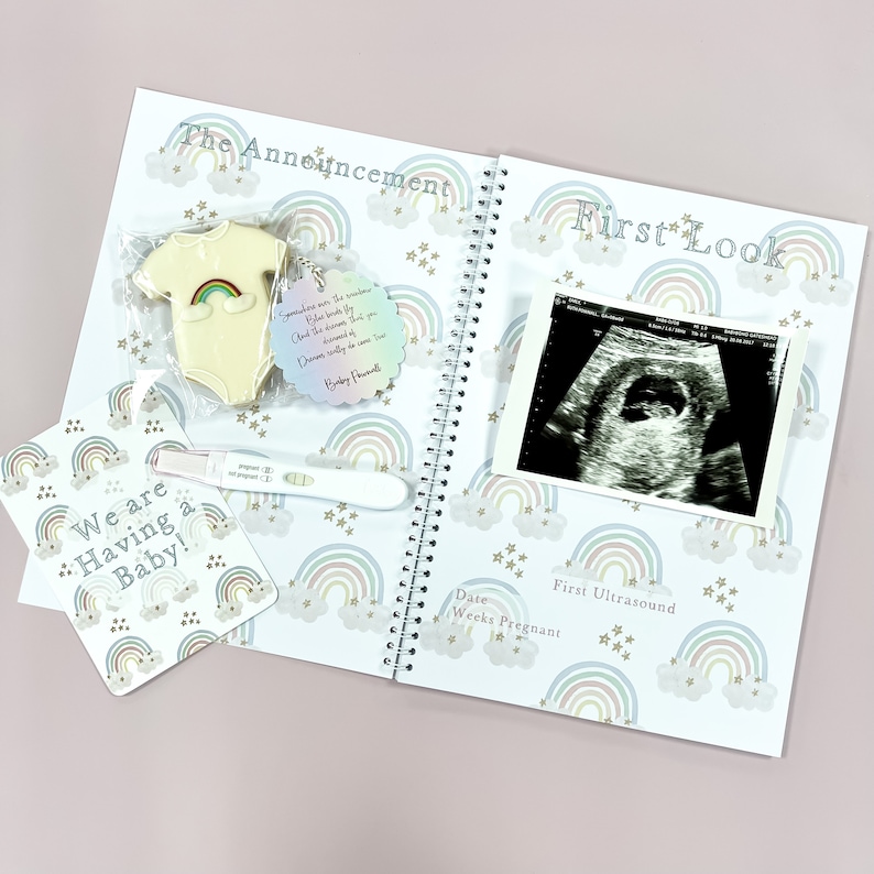 May include: A white baby onesie with a rainbow design is displayed on a white background with a rainbow and star pattern. The text "The Announcement" is at the top of the page. A pregnancy test with a positive result is visible in the lower left corner. A small tag with the text "Sometimes we are the rainbow.  You make the storm. And the heavens know you deserved every bit of the dreams we are now living." is attached to the onesie.