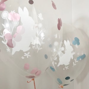 May include: Two clear balloons with white bunny silhouettes and confetti. The balloon on the left has pink confetti and the balloon on the right has blue confetti.