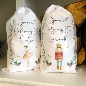 May include: Two white Christmas gift bags with personalized names. The bag on the left features a ballerina illustration and the text "Special Delivery For Edie". The bag on the right features a nutcracker illustration and the text "Special Delivery For Jacob".