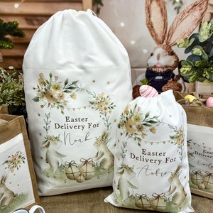 May include: Two white fabric Easter gift bags with bunny illustrations and floral designs. The bags have the text "Easter Delivery For" and personalized names "Noah" and "Aobie".