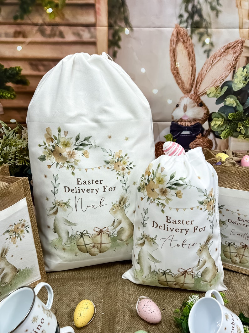 May include: Two white fabric Easter gift bags with bunny illustrations and floral designs. The bags have the text "Easter Delivery For" and personalized names "Noah" and "Aobie".