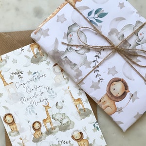 May include: A white card with a watercolor design of giraffes, lions, and elephants on clouds. The card says "Congratulations on your miracle in the making". The card is wrapped in white wrapping paper with a watercolor design of giraffes, lions, and elephants on clouds. The wrapping paper is tied with twine.