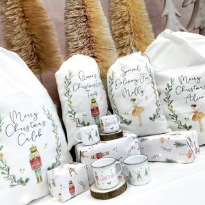 May include: Four white fabric gift bags with personalized Christmas messages and illustrations of a nutcracker and a ballerina. The bags are decorated with green leaves, red berries, and gold stars. There are also three small white mugs with a nutcracker design and the name "Jacob" on them.