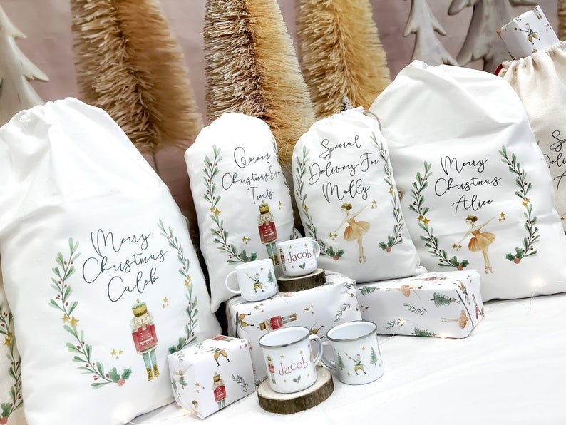 May include: Four white fabric gift bags with personalized Christmas messages and illustrations of a nutcracker and a ballerina. The bags are decorated with green leaves, red berries, and gold stars. There are also three small white mugs with a nutcracker design and the name "Jacob" on them.