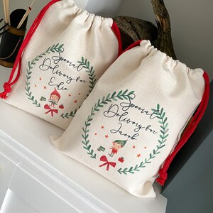 May include: Two white canvas gift bags with red drawstrings. Each bag has a green wreath with a Christmas elf and the text "Special Delivery for" followed by a name. The bag on the left says "Special Delivery for Eddie" and the bag on the right says "Special Delivery for Jacob".