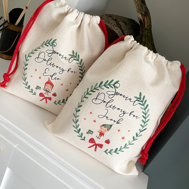 May include: Two white canvas gift bags with red drawstrings. Each bag has a green wreath with a Christmas elf and the text "Special Delivery for" followed by a name. The bag on the left says "Special Delivery for Eddie" and the bag on the right says "Special Delivery for Jacob".