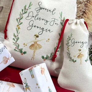 May include: Two white fabric gift bags with red drawstrings. The larger bag has a green wreath with gold stars and the text "Special Delivery for Georgia". The smaller bag has a similar wreath with the text "Georgia". Both bags feature a ballerina illustration.