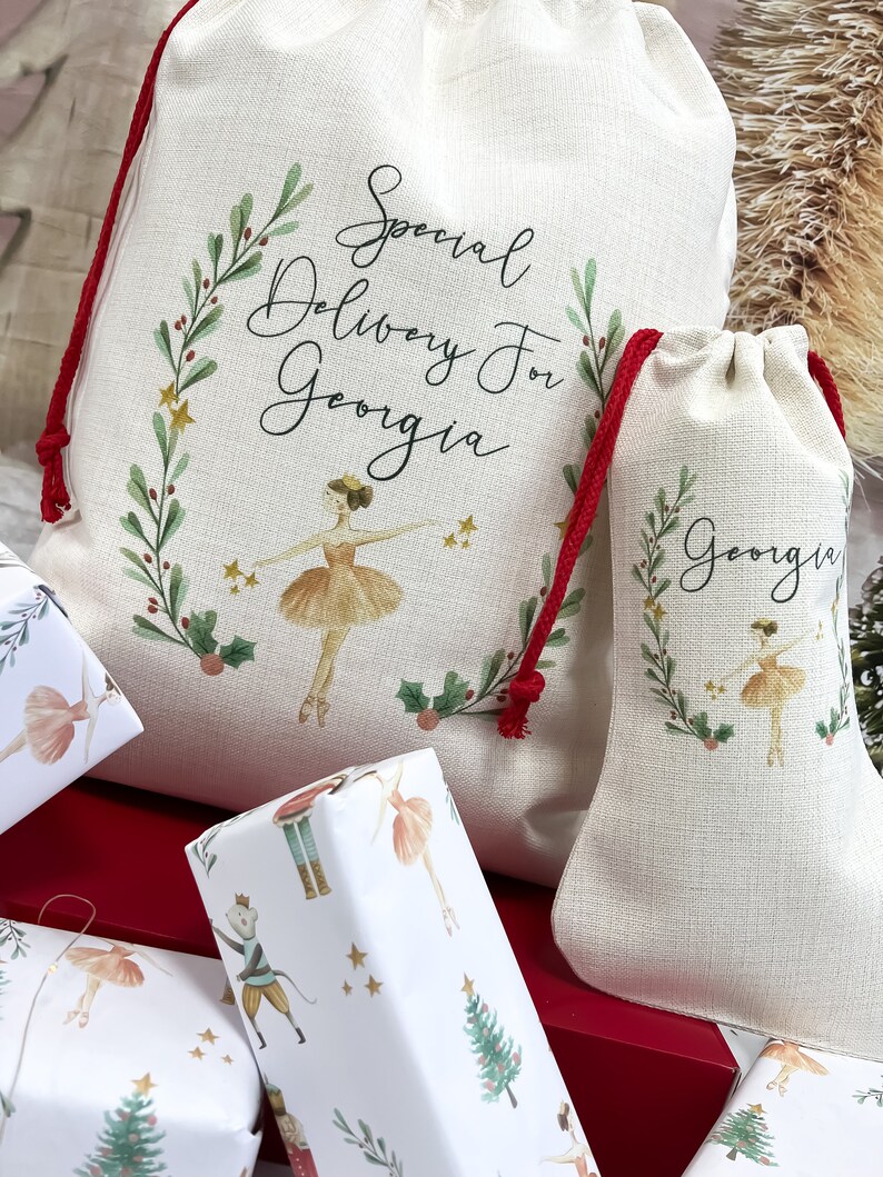 May include: Two white fabric gift bags with red drawstrings. The larger bag has a green wreath with gold stars and the text "Special Delivery for Georgia". The smaller bag has a similar wreath with the text "Georgia". Both bags feature a ballerina illustration.
