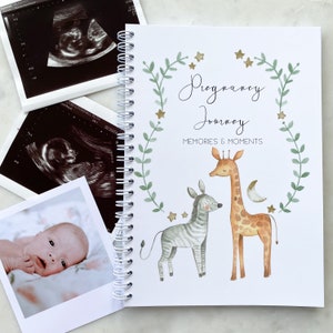 May include: A white pregnancy journal with a green leafy wreath and gold stars. The text "Pregnancy Journey" and "Memories & Moments" is printed on the cover. The journal features a watercolor illustration of a giraffe and zebra.
