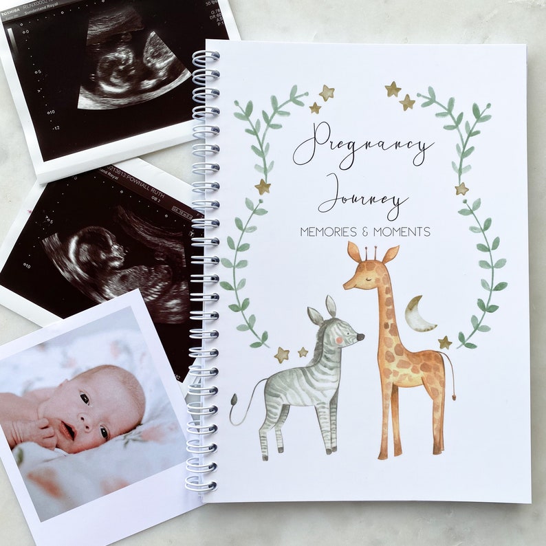 May include: A white pregnancy journal with a green leafy wreath and gold stars. The text "Pregnancy Journey" and "Memories & Moments" is printed on the cover. The journal features a watercolor illustration of a giraffe and zebra.