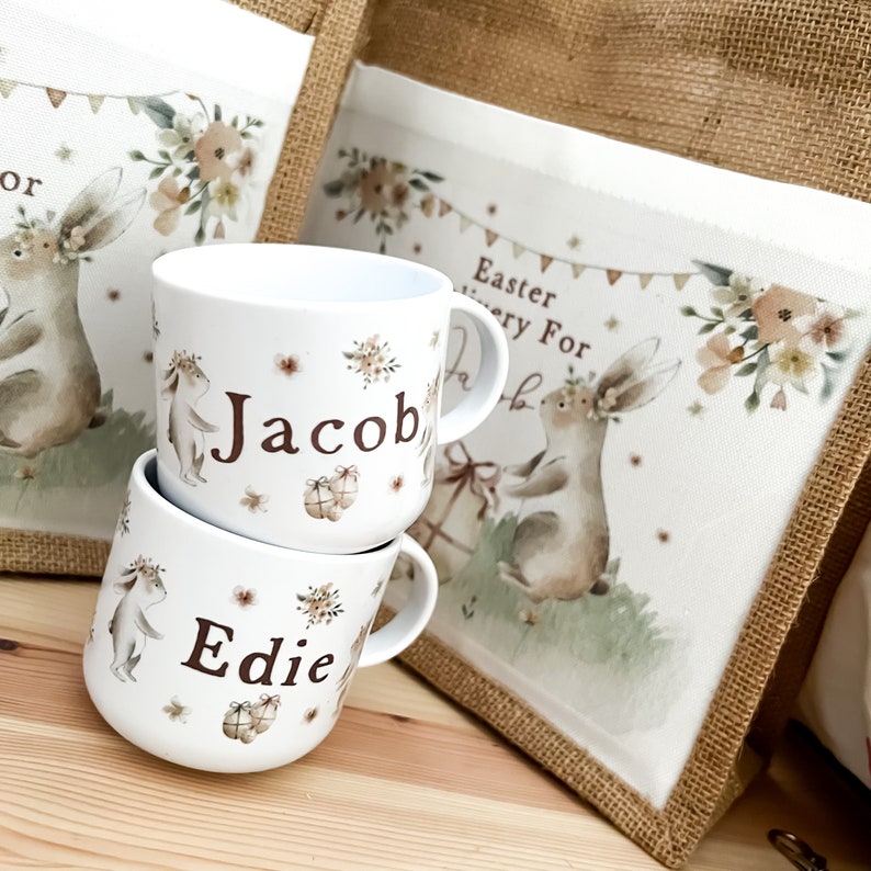 May include: Two white ceramic mugs with brown bunny and floral designs. The mugs are personalized with the names "Jacob" and "Edie". The mugs are sitting on a wooden surface in front of a burlap and white fabric bag with the text "Easter Every For" and a bunny design.