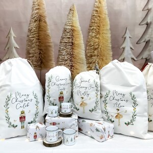 May include: Five white canvas gift bags with personalised Christmas messages and illustrations. The bags are decorated with green wreaths, gold stars, and images of a nutcracker, a ballerina, and a Christmas tree. The bags are arranged on a table with small gift boxes and mugs.