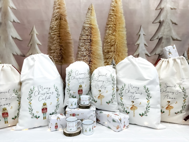 May include: Five white canvas gift bags with personalized Christmas messages and illustrations. The bags are decorated with green wreaths, gold stars, and images of a nutcracker, a ballerina, and a Christmas tree. The bags are arranged on a table with small gift boxes and mugs.