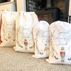 May include: Four white fabric gift bags with drawstring closures. Each bag features a different design with a festive wreath, a nutcracker, and a ballerina. The bags are personalized with the text "Special Delivery For" followed by a name.