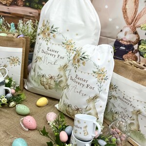 May include: Three white fabric Easter gift bags with bunny illustrations and floral designs. The bags are personalized with the names "Noah", "Aubrie", and "Emily". The bags are surrounded by pastel-colored Easter eggs and a small glass jar filled with chocolate eggs.