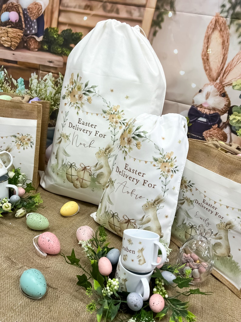 May include: Three white fabric Easter gift bags with bunny illustrations and floral designs. The bags are personalized with the names "Noah", "Aubrie", and "Emily". The bags are surrounded by pastel-colored Easter eggs and a small glass jar filled with chocolate eggs.