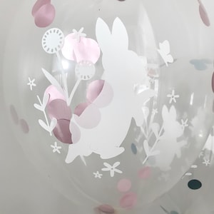May include: Clear balloon with a white bunny silhouette and pink confetti. The bunny is surrounded by white flowers and leaves.