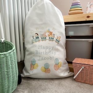 May include: White canvas toy bag with a colourful train design and the text "Happy Birthday Jacob!" printed on it. The train is filled with animals and has balloons above it.