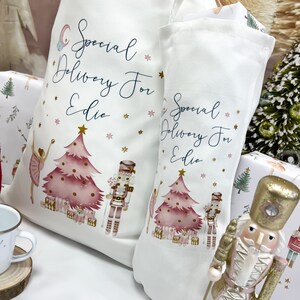 May include: Two white canvas stockings with pink and gold Christmas tree designs and the text "Special Delivery For Edie". A nutcracker figurine is in the foreground.