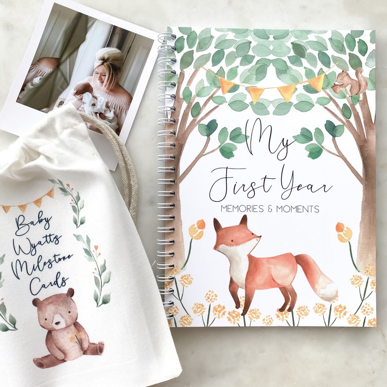 K&ouml;nnte beinhalten: Eine wei&szlig;e Stofftasche mit einer Illustration eines braunen B&auml;ren und dem Text "Baby Wyatt Milestone Cards". Ein spiralgebundenes Buch mit einer Aquarell-Illustration einer Waldszene, einem Fuchs und dem Text "My First Year Memories & Moments".