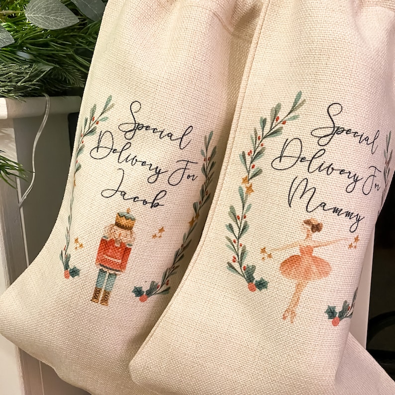 Pode incluir: Duas meias de Natal bege com um design festivo. Uma meia apresenta um quebra-nozes e o texto "Special Delivery For Jacob". A outra meia apresenta uma bailarina e o texto "Special Delivery For Mammy".