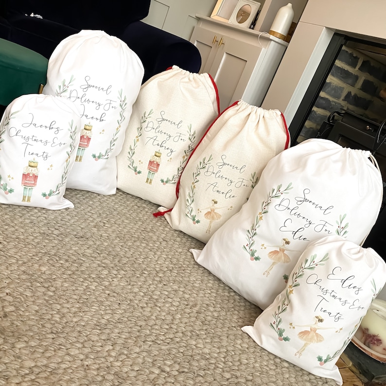 May include: Five white fabric gift bags with red drawstrings. Each bag has a different design featuring a watercolor illustration of a nutcracker or a ballerina. The bags are personalized with the names "Jacob", "Abbey", "Amelia", "Edie", and "Edie's Christmas Eve Treats".