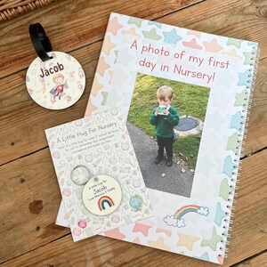 May include: A white and pink notebook with a colorful star pattern and the text "A photo of my first day in Nursery!"  A photo of a young boy in a green shirt and black pants is in the center of the notebook. A small white card with a rainbow and the text "A little hug for Jacob Love Mommy & Daddy" is in the bottom left corner. A round tag with a boy in a red cape and the text "Jacob" is in the top left corner.