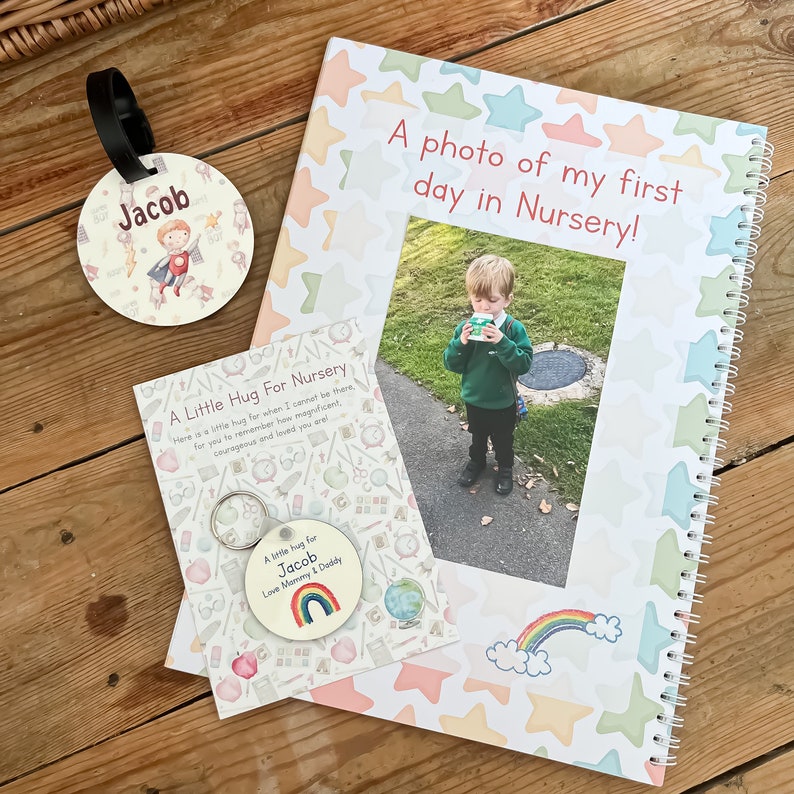 May include: A white and pink notebook with a colorful star pattern and the text "A photo of my first day in Nursery!"  A photo of a young boy in a green shirt and black pants is in the center of the notebook. A small white card with a rainbow and the text "A little hug for Jacob Love Mommy & Daddy" is in the bottom left corner. A round tag with a boy in a red cape and the text "Jacob" is in the top left corner.