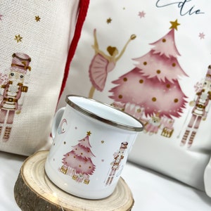 May include: A white enamel mug with a pink Christmas tree, a nutcracker, and stars. The mug is sitting on a wooden coaster.