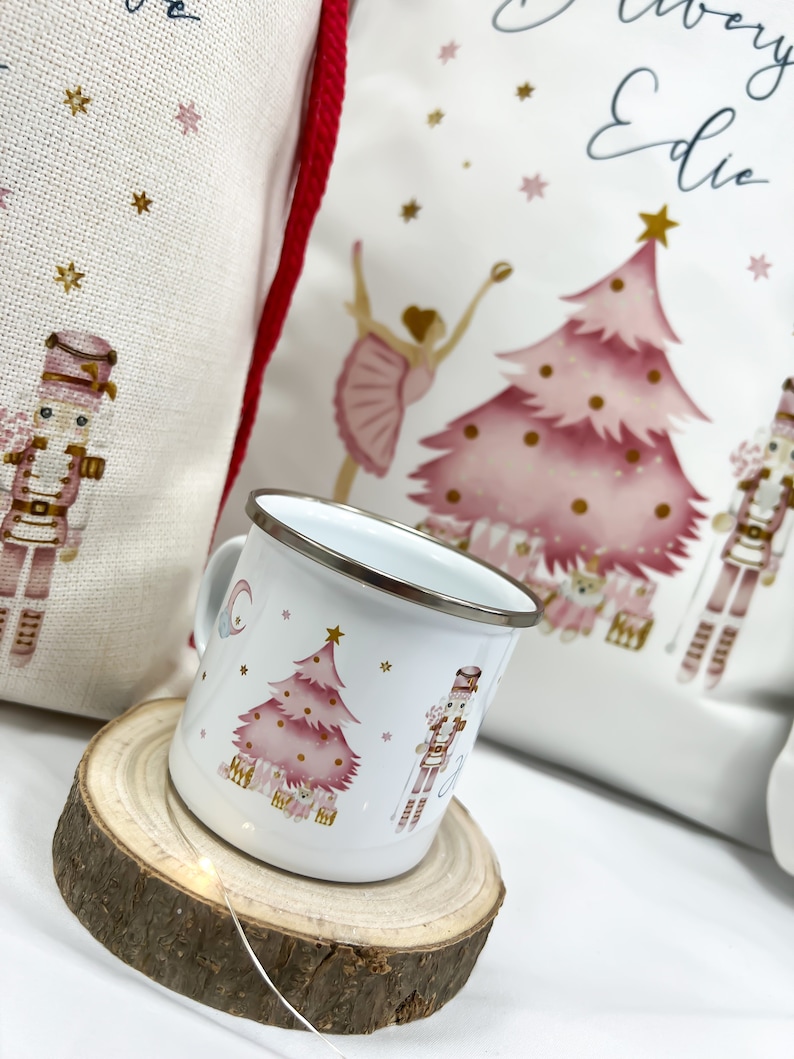 May include: A white enamel mug with a pink Christmas tree, a nutcracker, and stars. The mug is sitting on a wooden coaster.