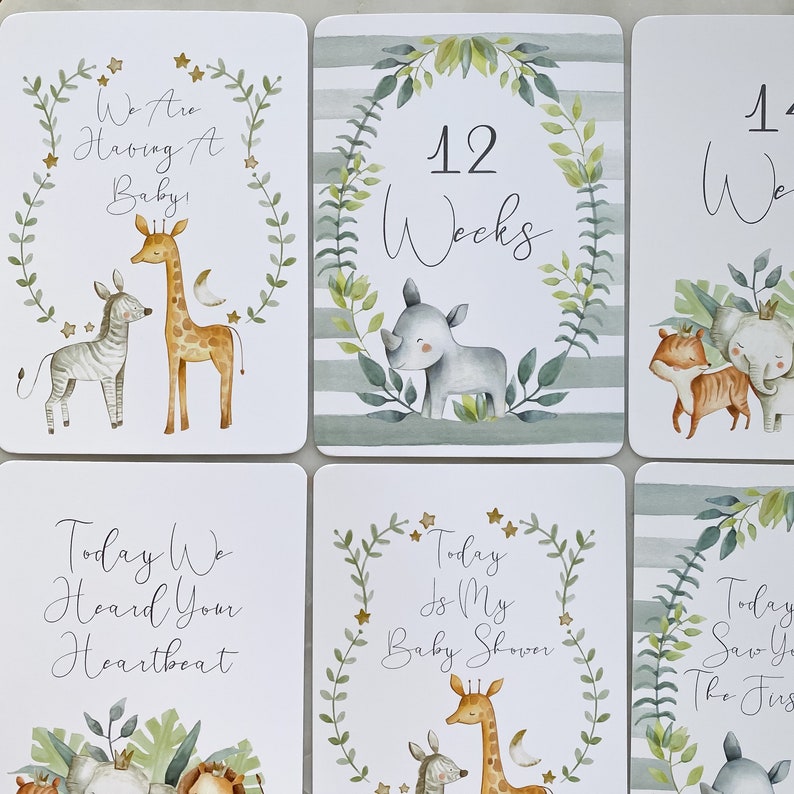 May include: A set of 8 baby shower milestone cards with jungle animal illustrations. The cards feature a giraffe, zebra, rhino, and elephant. The text on the cards includes "We are having a baby", "12 weeks", "Today we heard your heartbeat", "Today is my baby shower", and "Today saw you the first time".