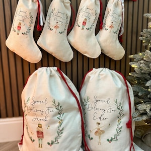 May include: Five personalized Christmas gift bags with red drawstrings. The bags are made of natural canvas and feature a festive design with a wreath, stars, and a choice of a nutcracker or ballerina. The bags are labeled with the names "Jacob", "Mommy", "Daddy", "Ellie", and "Edie".